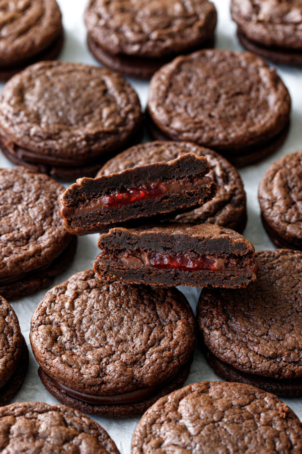 Staggered Chocolate Raspberry Sandwich Cookies, one cookie cut in half sitting on top to show the ganache and raspberry fillings.