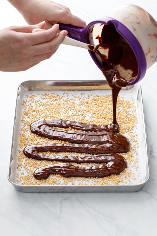 Pouring melted chocolate onto a parchment-lined pan with puffed quinoa scattered on top