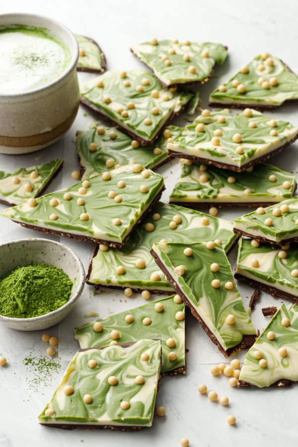 Uneven cut pieces of Matcha White Chocolate Crunch Bark, with small bowl of matcha tea powder and a cup of matcha latte in the background
