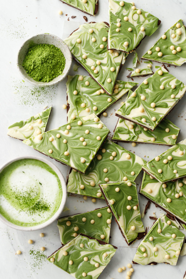 Overhead, scattered cut pieces of Matcha White Chocolate Crunch Bark, with bowl of matcha powder and a cup of frothed matcha latte