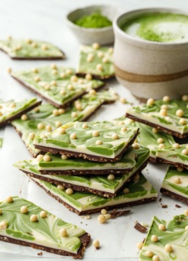 Stack of cut pieces of Matcha White Chocolate Crunch Bark showing the layers, cup with matcha latte and bowl of matcha powder in the background