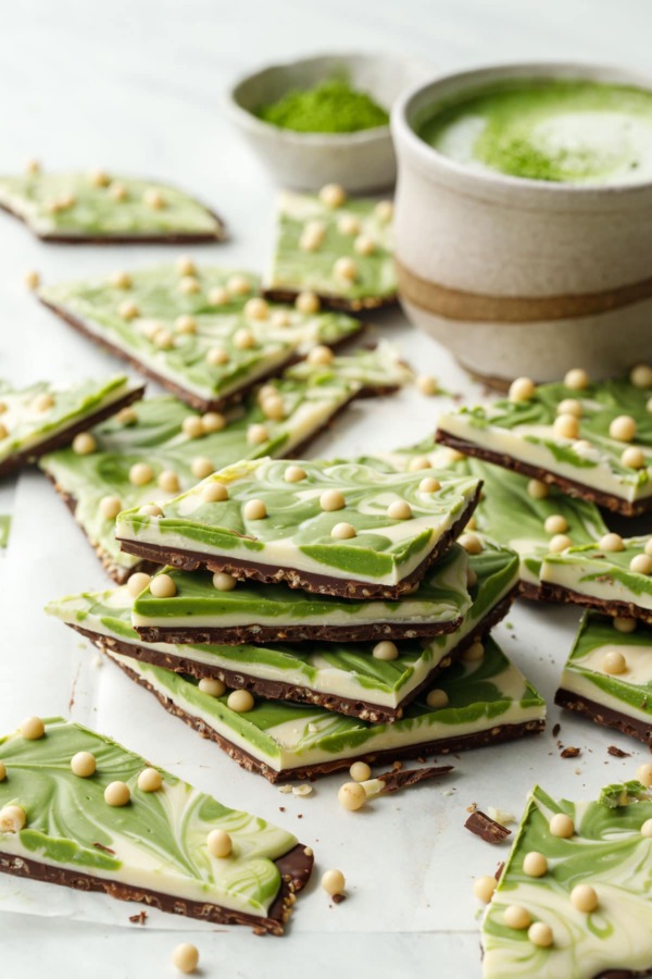 Stack of cut pieces of Matcha White Chocolate Crunch Bark showing the layers, cup with matcha latte and bowl of matcha powder in the background