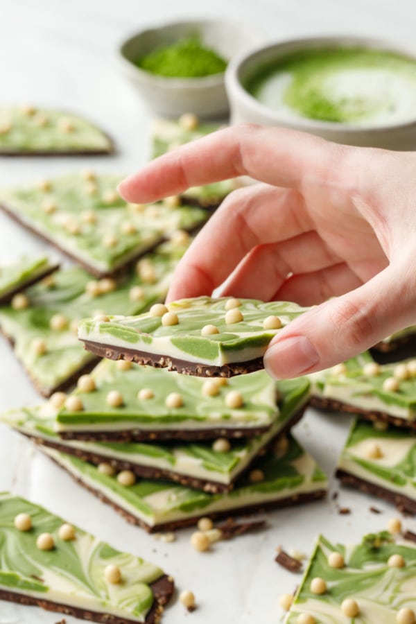 Hand lifting a cut piece of Matcha White Chocolate Crunch Bark, at an angle to show the dark and white chocolate layers