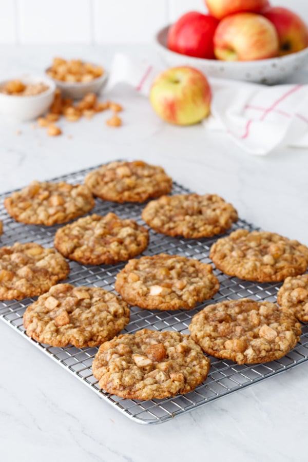 Wire rack with rows of Toffee Apple Oatmeal Cookies, bowl of apples and scattered cookie mix-ins in the background.