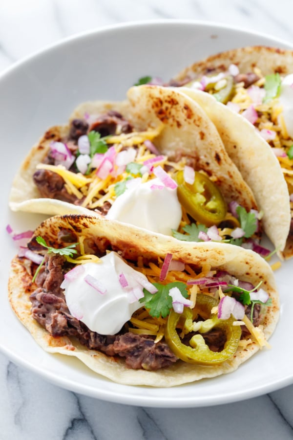 Three folded Easy Refried Black Bean Tacos on a white plate, with big dollops of sour cream and other toppings