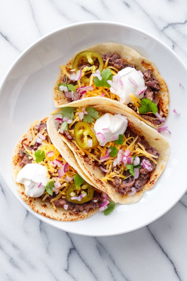 Overhead, three Easy Refried Black Bean Tacos on a white plate, white marble background