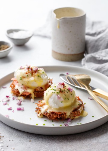 White plate with two Latke Eggs Benedicts and gold utensils, pitcher of hollandaise sauce in the background