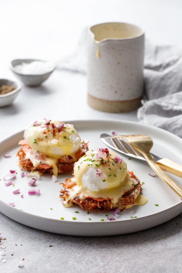 White plate with two Latke Eggs Benedicts and gold utensils, pitcher of hollandaise sauce in the background