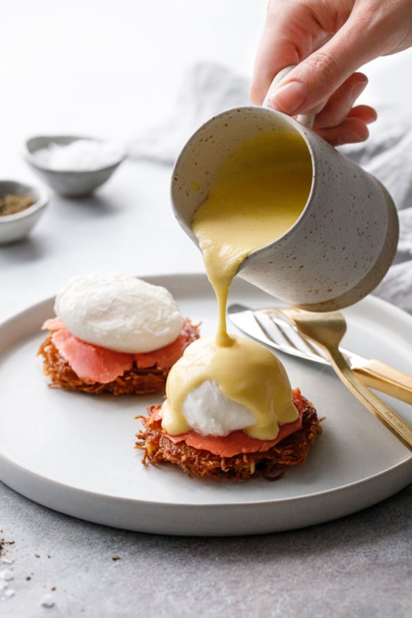 Pouring hollandaise sauce on top of a perfectly poached egg, smoked salmon, and crispy potato latke