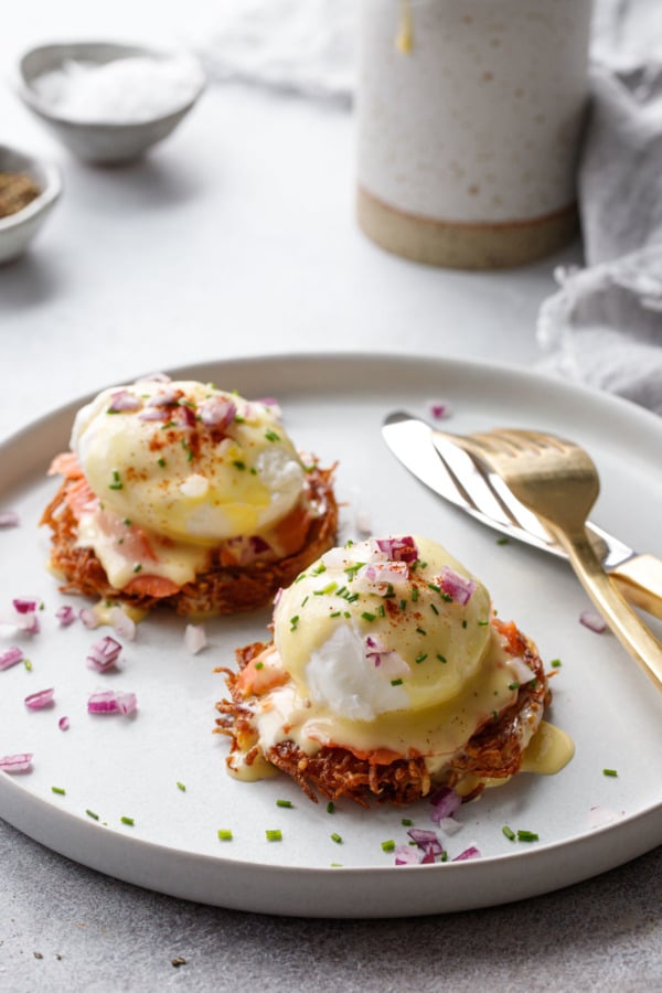 Latke Eggs Benedict, garnished with chives and red onion, on a white plate, gold utensils.