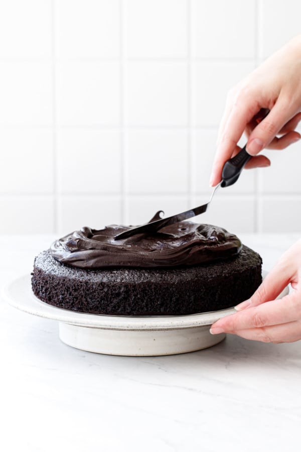 Нанесение глянцевой шоколадной глазури на сметанный торт из темного шоколада на белой подставке для торта.