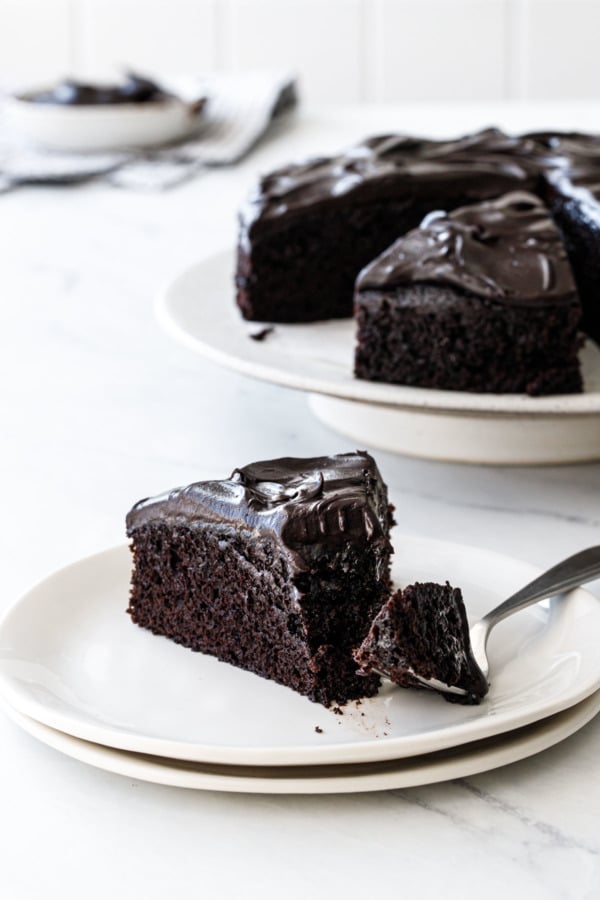 Plate with a cut slice of Sour Cream Chocolate Cake and a forkful taken out of it, full cake on a cake stand in the background