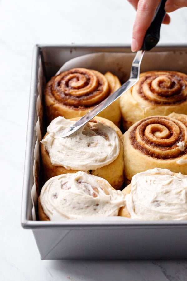 Spreading brown butter cream cheese frosting on a pan of freshly baked cinnamon rolls