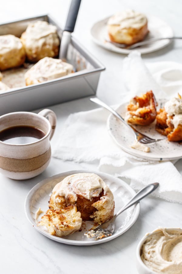 Brown Butter Cinnamon Rolls, one partially eaten on a plate in the foreground, coffee, more plates, and baking pan in the background