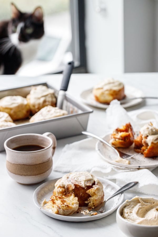 Pull back of photo scene with multiple plates and pan of cinnamon rolls, with a cat out of focus in the background.