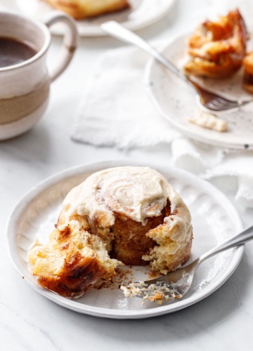 One Brown Butter Cinnamon Roll on a ceramic plate, with a forkful to show the pillowy texture.