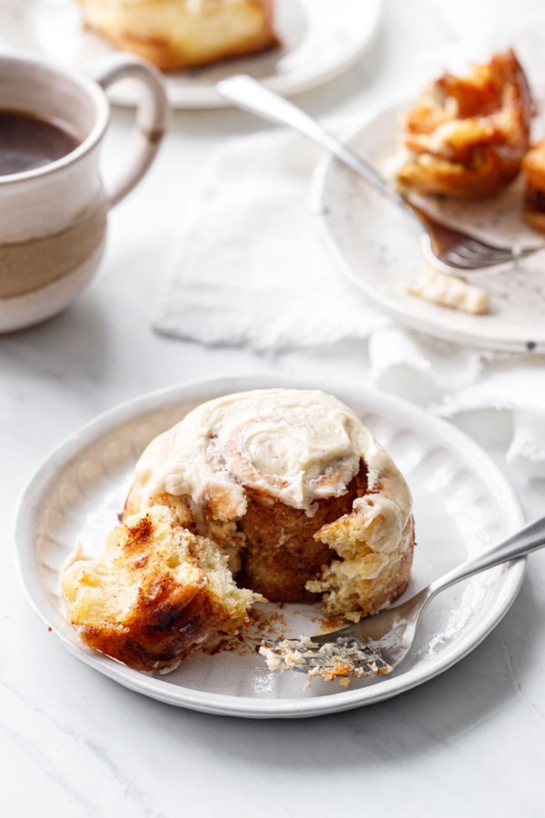 One Brown Butter Cinnamon Roll on a ceramic plate, with a forkful to show the pillowy texture.