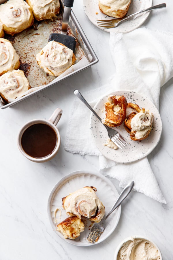Overhead, plates of Brown Butter Cinnamon Roll in various states of consumption, with pan of rolls and a cup of coffee on a white marble background