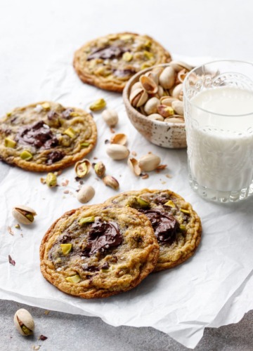 A few Salted Pistachio & Dark Chocolate Chunk Cookies on a piece of crinkled parchment, with a glass of milk and bowl of shelled pistachios.