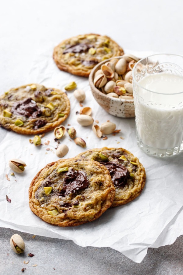 A few Salted Pistachio & Dark Chocolate Chunk Cookies on a piece of crinkled parchment, with a glass of milk and bowl of shelled pistachios.