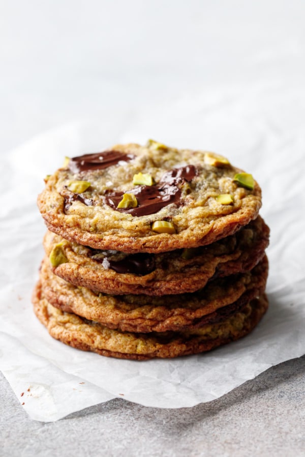 Stack of four Salted Pistachio & Dark Chocolate Chunk Cookies on crinkled parchment, puddles of melted chocolate reflecting the light.