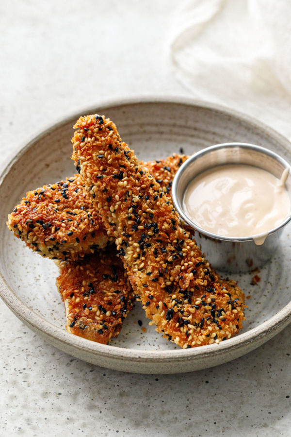 Three Panko Sesame Chicken Tenders in a ceramic dish with a small metal container of sriracha mayo