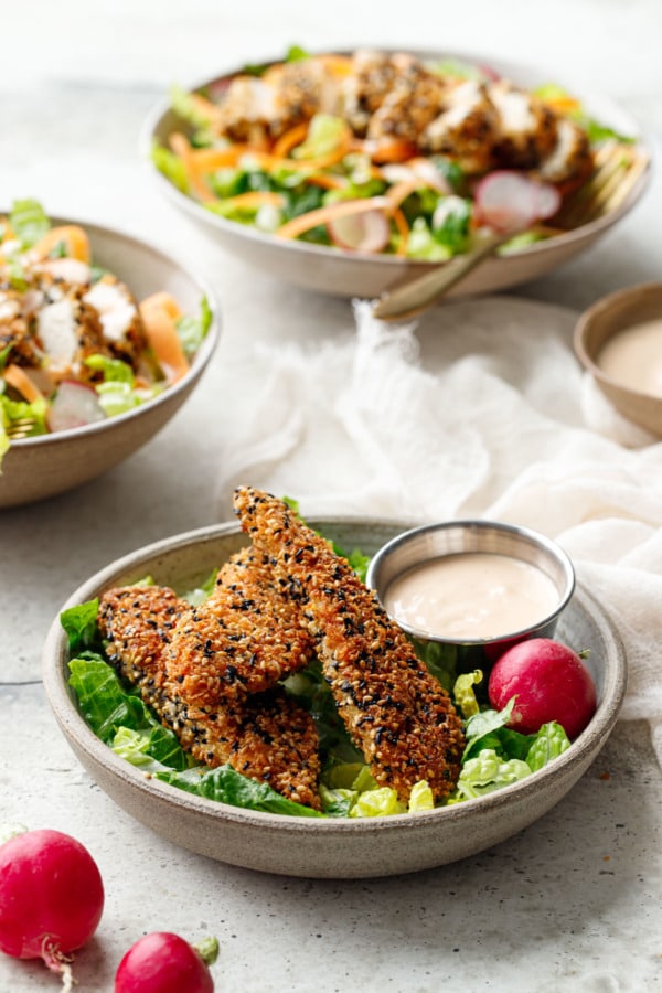 Panko Sesame Chicken Tenders in a bowl with lettuce and dipping sauce, bowls of salad topped with cut up tenders in the background