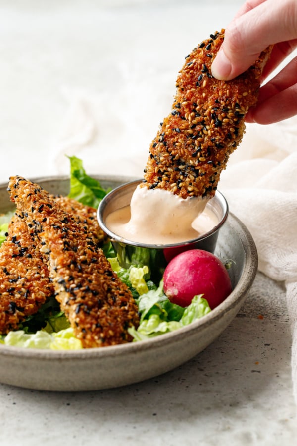 Dipping a Panko Sesame Chicken Tender into a metal dip container with sriracha mayo, lettuce and radish on the side