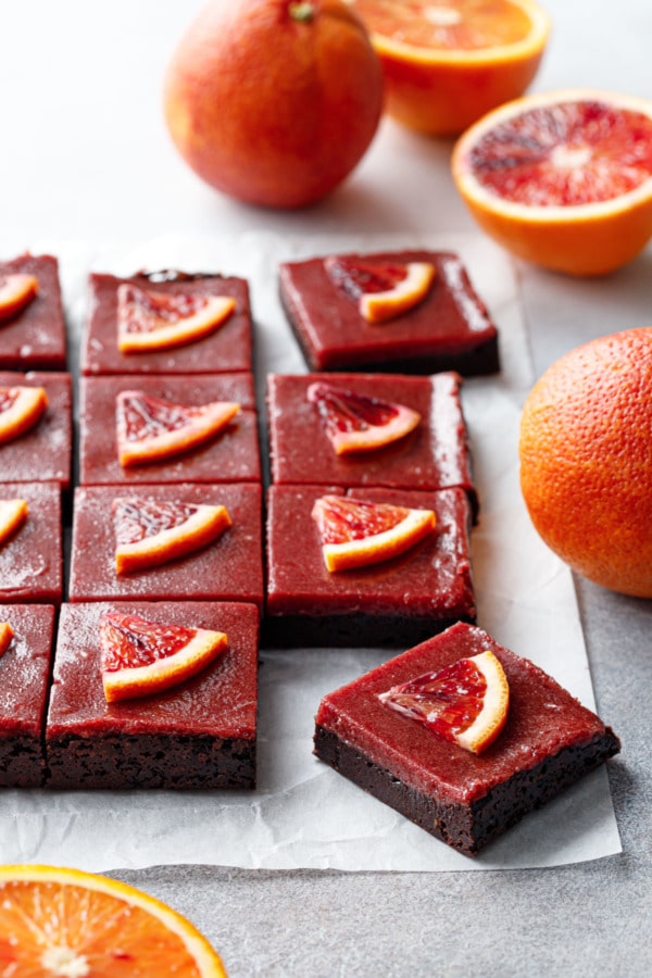 Cut squares of Blood Orange Curd Brownies topped wtih little wedges of blood orange, on parchment with whole and cut oranges around.