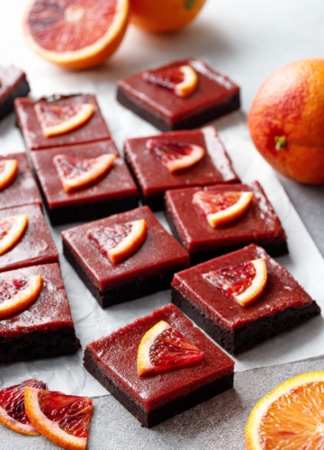 Cut squares of Blood Orange Curd Brownies on parchment, with orange slices and whole and halved oranges scattered around.