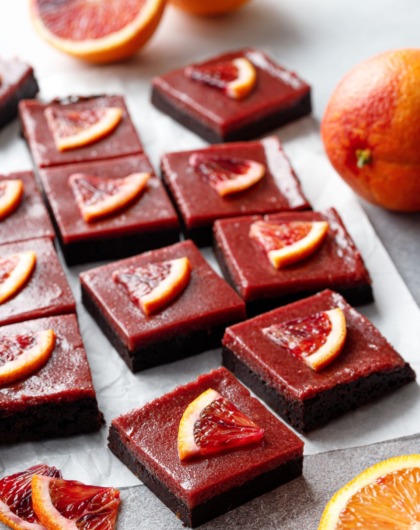 Cut squares of Blood Orange Curd Brownies on parchment, with orange slices and whole and halved oranges scattered around.