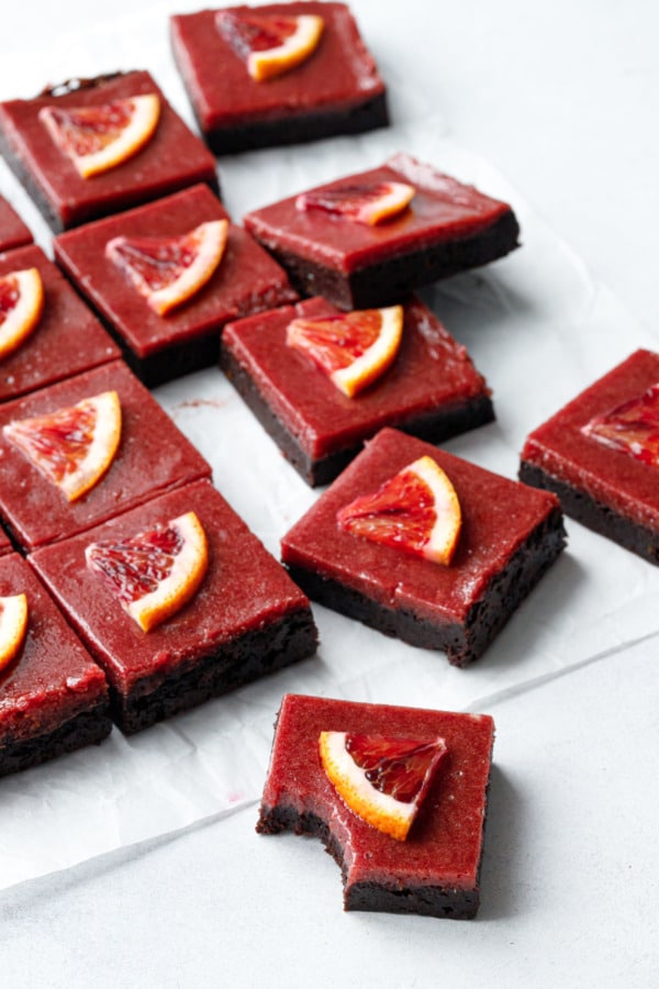 Messily arranged squares of Blood Orange Curd Brownies on a white parchment, one piece with a bite out of it