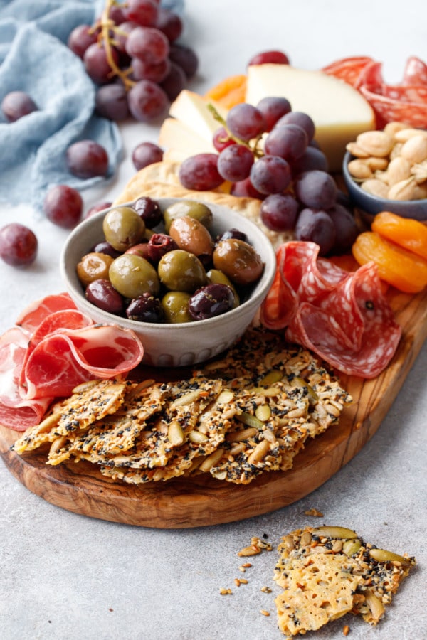 Olivewood cheeseboard with all the fixings, fan of Seeds and Cheese Crackers in the front and a bowl of olives, charcuterie, and grapes