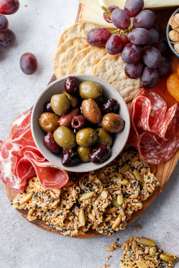 Overhead, cheeseboard with all the fixings, fan of Seeds and Cheese Crackers in the front and a bowl of olives, charcuterie, and grapes
