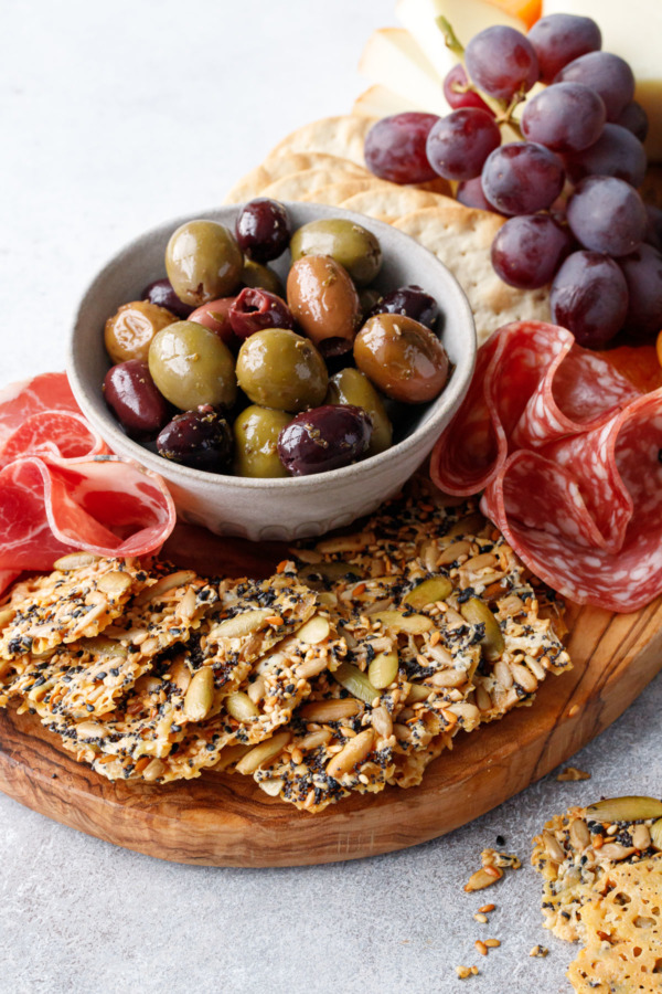 Olivewood cheeseboard with fan of Seeds and Cheese Crackers in the front and a bowl of olives, charcuterie, and grapes
