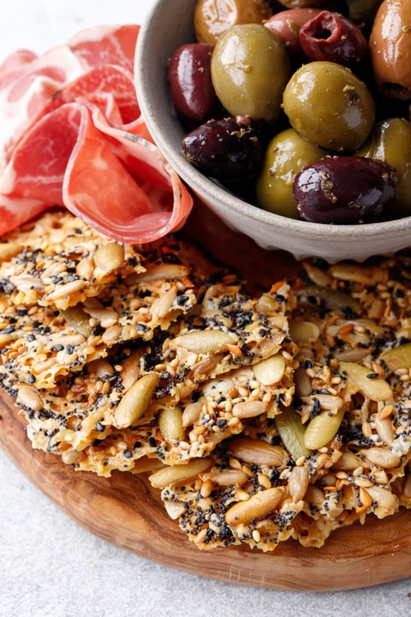 Closeup, olivewood cheeseboard with fan of Seeds and Cheese Crackers and a bowl of olives and charcuterie.