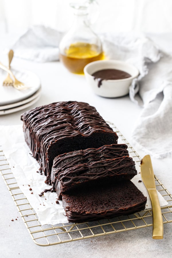 Sliced Chocolate Olive Oil Loaf Cake on a wire rack with gold knife, plates and bottle of olive oil in the background.