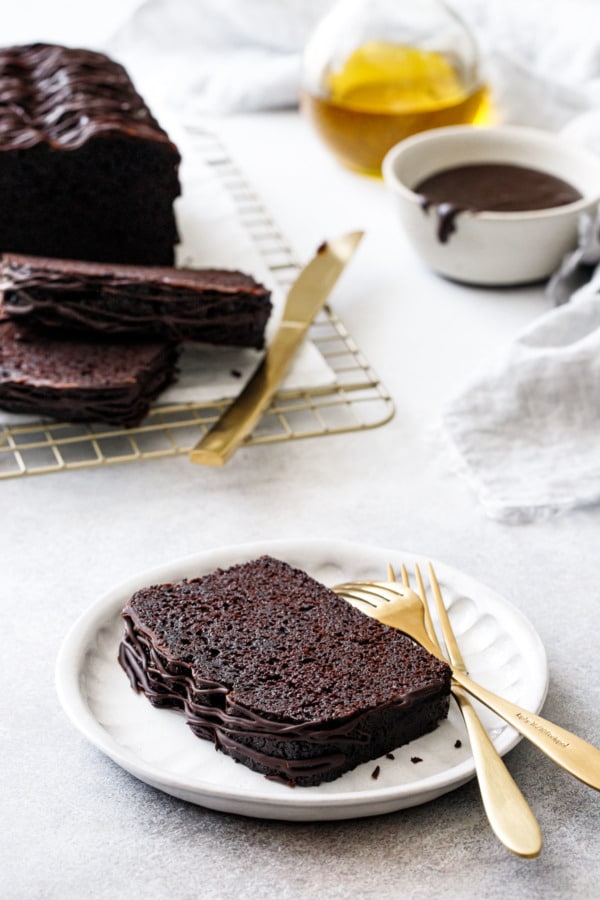 Slice of Chocolate Olive Oil Loaf Cake on a plate with two gold knives, partially sliced loaf in the background with bowl of glaze and bottle of olive oil.