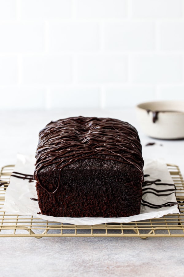 Freshly glazed Chocolate Olive Oil Loaf Cake on a wire rack, white subway tile background