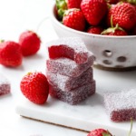 Stack of Homemade Strawberry Gummies coated in sour sugar, one candy with a bite out of it; fresh strawberries scattered around.