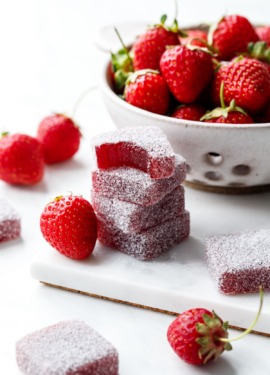 Stack of Homemade Strawberry Gummies coated in sour sugar, one candy with a bite out of it; fresh strawberries scattered around.