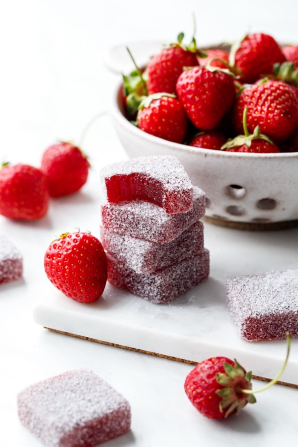 Stack of Homemade Strawberry Gummies coated in sour sugar, one candy with a bite out of it; fresh strawberries scattered around.