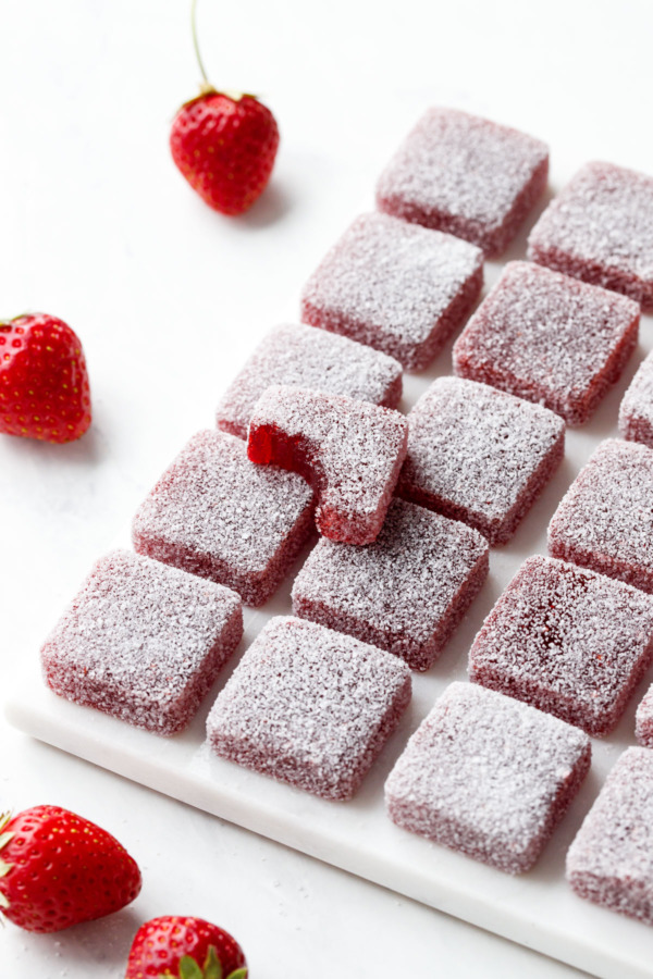 Grid of square Homemade Sour Strawberry Gummies on marble, one piece with a bite taken out of it on top.