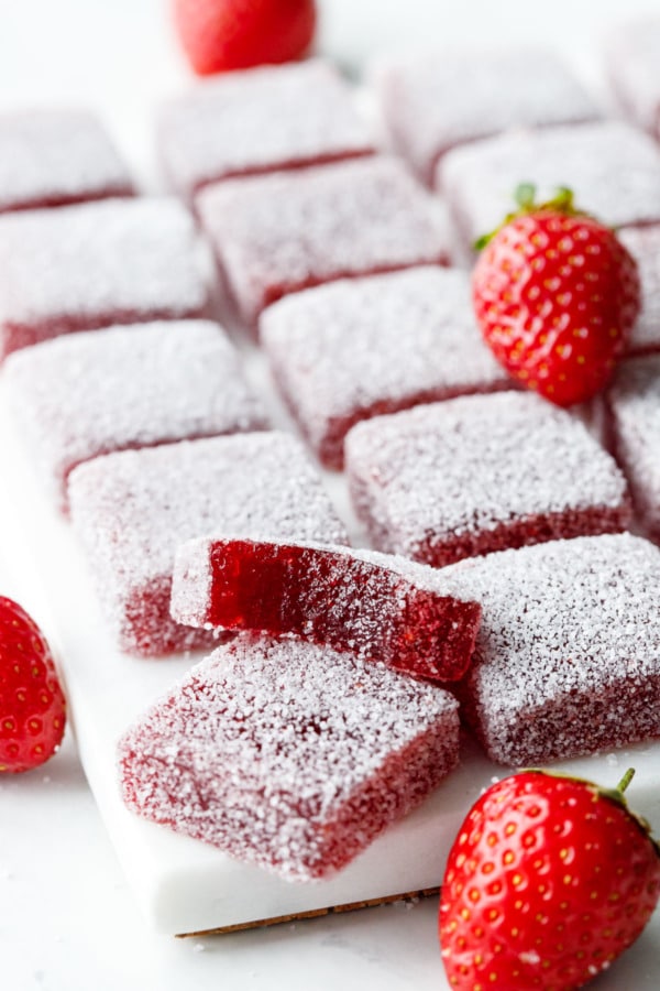 Grid of square Homemade Sour Strawberry Gummy candies, with fresh strawberries and one piece with a bite out of it.