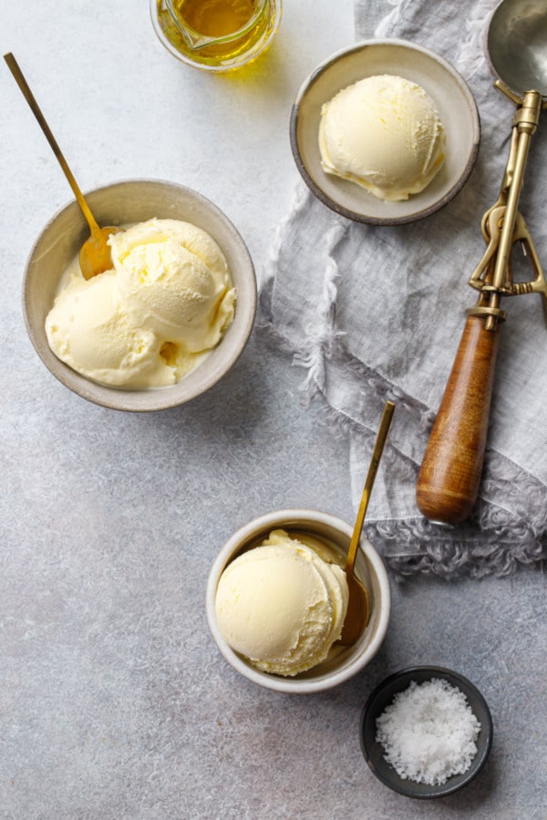 Overhead, three mis-matched bowls with scoops of olive oil ice cream, vintage ice cream scoop and tiny pitcher of olive oil