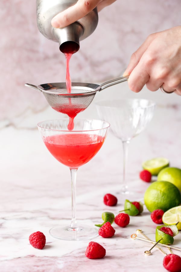 Hands pouring Raspberry Lime Gin Sour through a silver sieve into a faceted martini glass