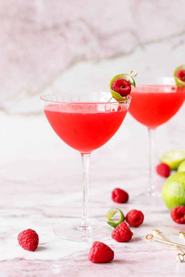 Two glasses with bright pink Raspberry Lime Gin Sours, garnished with lime peel and fresh raspberry on a pink marble background.