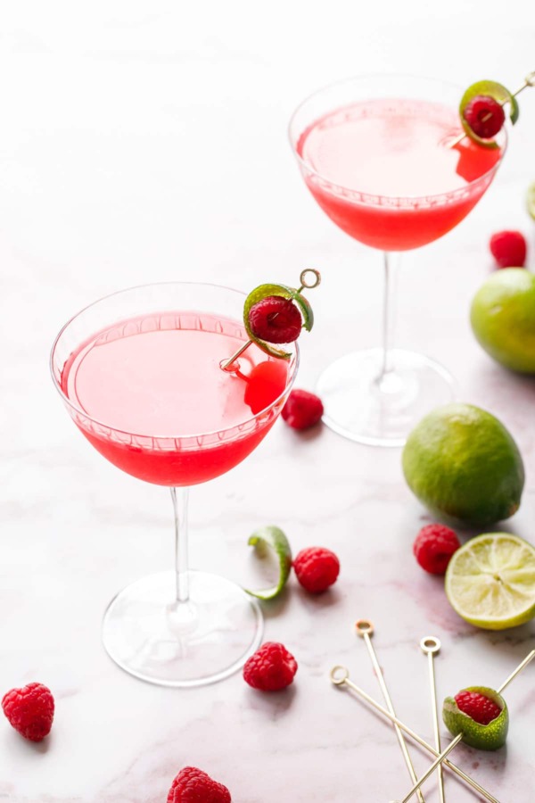 Backlit glasses with bright pink Raspberry Lime Gin Sours, garnished with lime peel and fresh raspberry on a pink marble background.