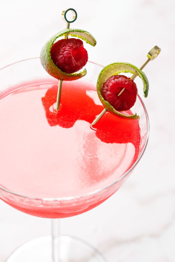 Closeup, backlit glass of Raspberry Lime Gin Sour, with two cocktail picks and lime raspberry garnish