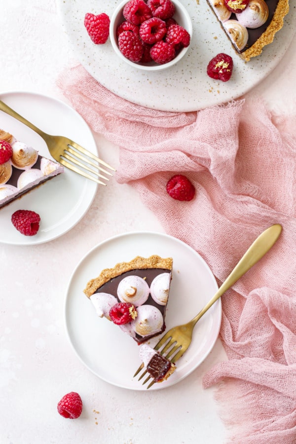 Overhead, two small dessert plates with triangular slices of Chocolate Raspberry S'mores Tart, pink background and pink linen with fresh raspberries scattered around.